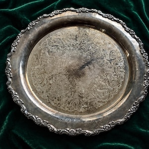 May include: A silver-colored, antique serving tray with an ornate floral and scroll design. The tray has a detailed, scalloped edge with small floral accents. The surface of the tray shows signs of wear, adding to its vintage appeal. The tray is set against a dark green velvet background.