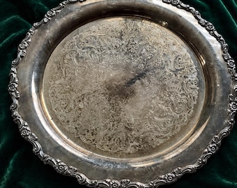 May include: A silver-colored, antique serving tray with an ornate floral and scroll design. The tray has a detailed, scalloped edge with small floral accents. The surface of the tray shows signs of wear, adding to its vintage appeal. The tray is set against a dark green velvet background.