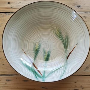 May include: A round, off-white ceramic bowl with a brown rim and a wheat stalk design. The bowl features concentric circle patterns and green and brown wheat illustrations. The bowl is set against a wooden surface.