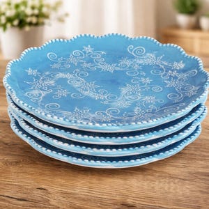 May include: A stack of four light blue ceramic plates with a scalloped edge and white beaded trim. Each plate features a white snowflake and swirl design. The plates are stacked on a wooden surface.