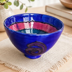 May include: A vibrant blue ceramic bowl with a decorative purple swirl design on the side. The interior features bands of pink and teal. The bowl sits on a beige linen placemat. A decorative piece for the kitchen or dining room.