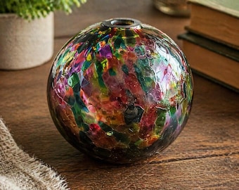 May include: A round, colorful glass vase with a textured surface. The vase features a mosaic of red, green, purple, and yellow hues. The top of the vase has a small opening. The vase is sitting on a wooden surface.