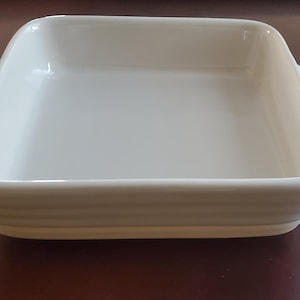 May include: A square, off-white ceramic baking dish with two curved handles. The dish has a smooth interior and a ribbed exterior, perfect for baking casseroles or side dishes. The dish is sitting on a dark brown surface.