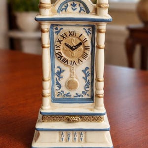 May include: A decorative ceramic clock in a rectangular shape. The clock features a cream-colored base with blue and gold accents. The clock face has Roman numerals and black clock hands. The clock is approximately 6 inches tall.