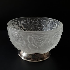 May include: A clear glass bowl with a frosted rose design, set on a silver-colored base. The bowl's exterior features detailed rose carvings, creating a textured surface. The bowl is likely used for decorative purposes.