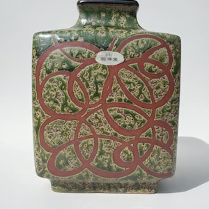May include: A square ceramic vase with a textured green and brown surface. The vase features a red, interconnected, circular design on the front. A small white label with Japanese text is affixed to the front of the vase.