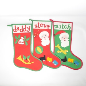May include: Three felt Christmas stockings with personalized names: "daddy", "steve", and "mitch". Each stocking features a different festive design, including a bell, a Santa Claus, and a toy airplane.