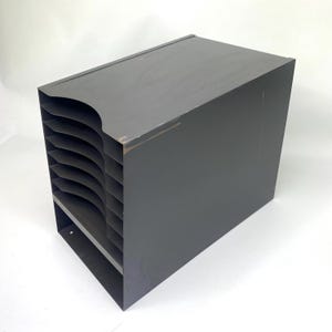 May include: A dark grey metal desk organiser featuring a vertical design with six stacked horizontal compartments for document storage. The unit has a solid rectangular frame with a curved cutout on the front edge of each shelf for easy access.