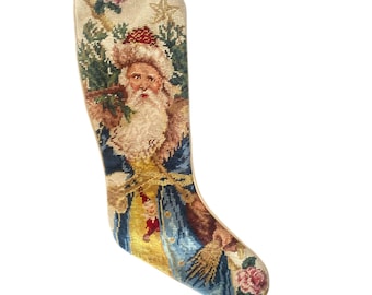 Puede incluir: Un calcetín navideño de punto de aguja con una representación detallada de Santa Claus con gorro rojo y abrigo azul con ribetes dorados. El diseño incluye una barba blanca, una pequeña figura de elfo, una estrella y flores rosas sobre fondo crema.