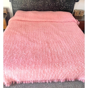 May include: A soft, light pink blanket on a bed. The blanket has a shaggy texture and a fringe along the bottom edge. The bed is against a dark, patterned headboard.