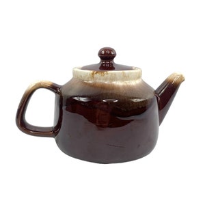 Puede incluir: Una tetera de cerámica esmaltada marrón y blanca con cuerpo redondeado y un pico. La tetera tiene un esmalte marrón con una banda blanca alrededor de la parte superior.