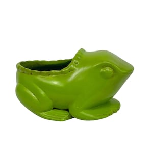 Puede incluir: Una maceta verde brillante con forma de rana. La maceta tiene una amplia abertura para plantas y está diseñada con características de rana, incluyendo ojos, patas y un borde texturizado. La maceta está hecha de un material sólido y es adecuada para uso en interiores o exteriores.
