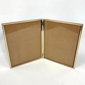May include: A gold-toned, hinged double picture frame. Each side has a rectangular opening with a gold border and a cardboard backing. The frame is open, displaying the cardboard backing. The frame is on a white surface.