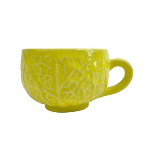 May include: A bright yellow ceramic mug with a cabbage leaf design. The mug has a rounded shape and a handle.
