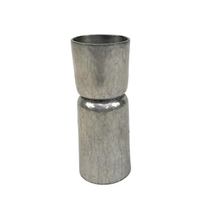 May include: A silver-coloured, two-sided jigger with a hammered texture. The jigger has a cylindrical shape with a smaller cup on top and a larger cup below, connected by a rounded middle section. The metal has a slightly aged appearance.