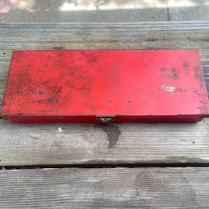 May include: A rectangular red metal storage box featuring a hinged lid and a single front latch. The exterior surface shows signs of wear, including various scratches, scuffs, and areas of paint loss that reveal the underlying metal texture.
