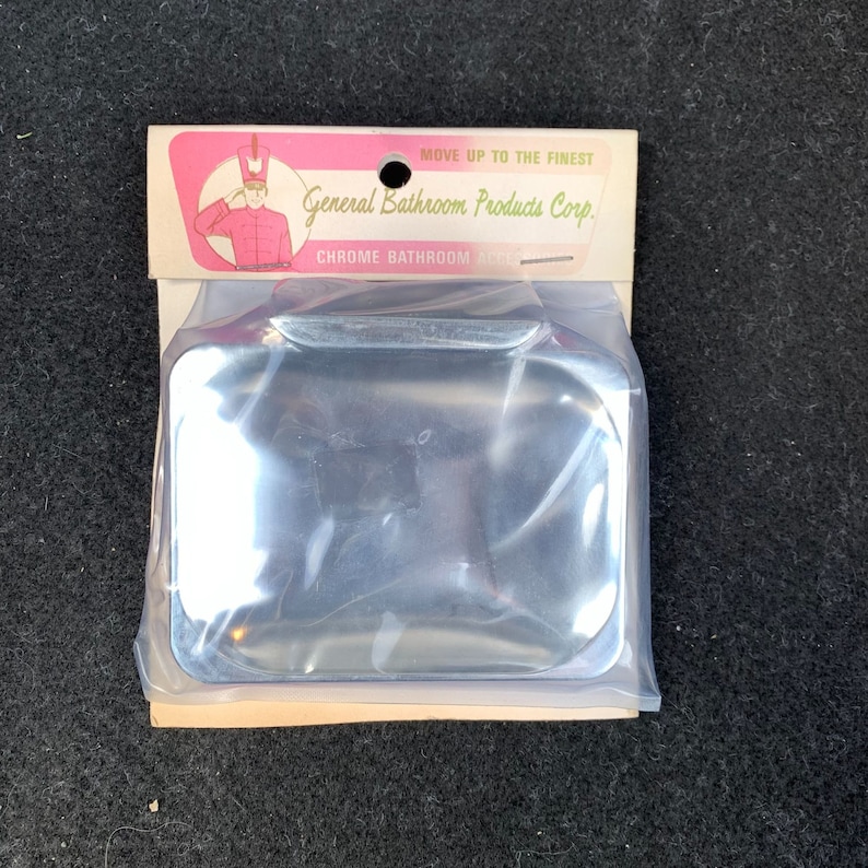 May include: A chrome bathroom accessory, square-shaped, packaged in clear plastic. The packaging includes a cardboard header with the text "General Bathroom Products Corp." and an illustration of a person in a red uniform saluting.