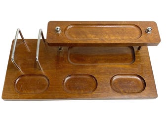 May include: A vintage wooden desk organizer with a rich brown finish. It features a raised top section with a long, oval indentation, three smaller oval indentations, and two silver metal pen holders. The organizer is designed to hold various desk supplies.