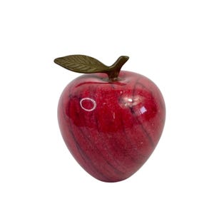 Könnte beinhalten: Ein dekorativer roter Marmorapfel mit dunkelroten Adern. Der Apfel hat einen bronzefarbenen Stiel und ein Blatt. Die polierte Oberfläche reflektiert das Licht und erzeugt Glanzlichter. Diese fruchtförmige Skulptur ist ein einzigartiger Einrichtungsgegenstand.