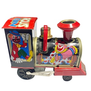 Pode incluir: Uma locomotiva de comboio de circo de brinquedo em folha de flandres vintage com um telhado vermelho com o texto CIRCUS TRAIN, corpo preto com ilustrações de palhaços, um sino dourado, uma chaminé vermelha e rodas pretas com biela de metal prateado.