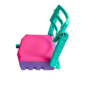 Puede incluir: Una silla plegable de color turquesa y morado con un asiento rosa y las palabras "COOL GEAR" impresas en la parte posterior.