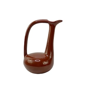 Puede incluir: Una jarra de cerámica roja con un asa curva única y un cuerpo redondeado. La jarra está esmaltada en un color rojo intenso y rico.
