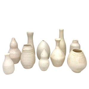 Pode incluir: Uma coleção de dez vasos de cerâmica branco sujo em várias formas e tamanhos. Alguns vasos são lisos, enquanto outros têm superfícies texturizadas ou desenhos canelados. Os vasos estão dispostos sobre um fundo branco.