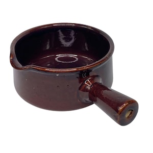 May include: A small, brown ceramic saucepan with a handle. The pan has a pouring spout and a glossy finish. The handle is attached to the side of the pan and has a hole at the end. The saucepan is designed for cooking and serving.