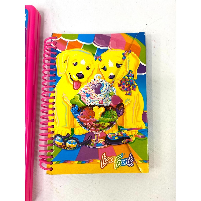 May include: A spiral-bound notebook with a colorful cover featuring two yellow cartoon dogs and a sundae. The notebook has a pink spiral binding and the words "Lisa Frank" are printed on the bottom right corner.
