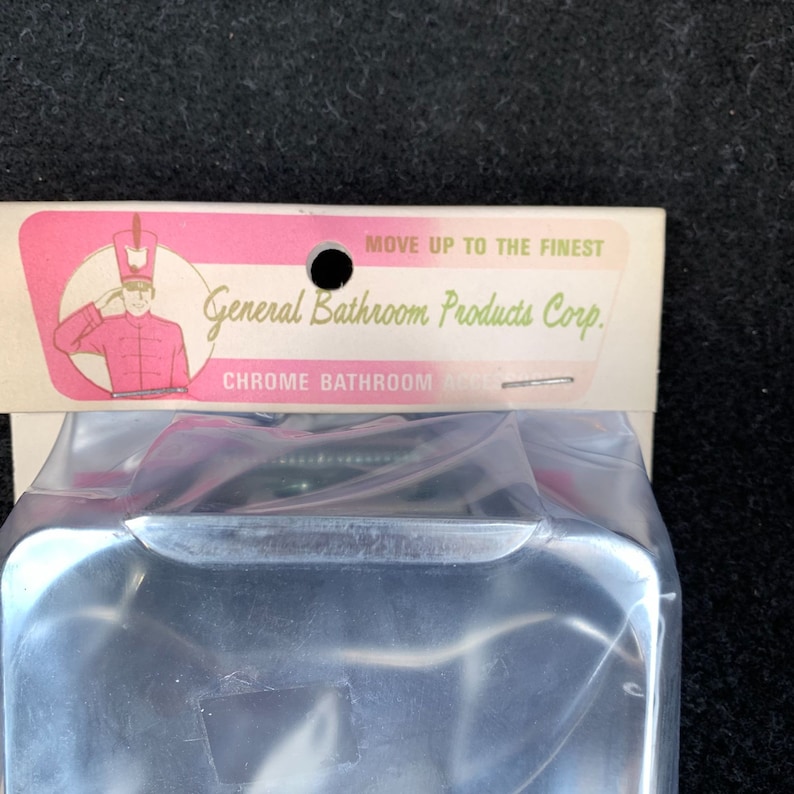 May include: A chrome bathroom accessory, partially visible, packaged in clear plastic. The packaging features a pink and white label with the text "MOVE UP TO THE FINEST" and "General Bathroom Products Corp."