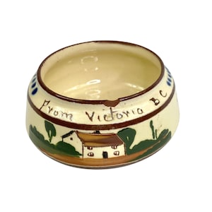 May include: Cream-colored ceramic ashtray with a brown rim and base. The ashtray features a hand-painted landscape scene with a house and the text "From Victoria BC". Blue and green accents complete the design.