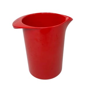 May include: A bright red plastic measuring cup with a handle and a pouring spout. The cup is cylindrical with a wide opening and a curved handle on one side. The spout is on the opposite side of the handle.