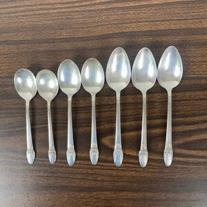 May include: Seven silver spoons with ornate handles. The spoons are arranged in a row on a wooden surface.