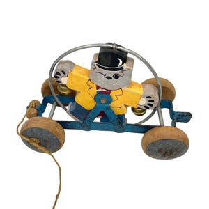 May include: A vintage wooden pull toy featuring a gray cat character wearing a yellow jacket and black hat. The cat is on a blue cart with wooden wheels and a metal hoop. A string is attached for pulling.