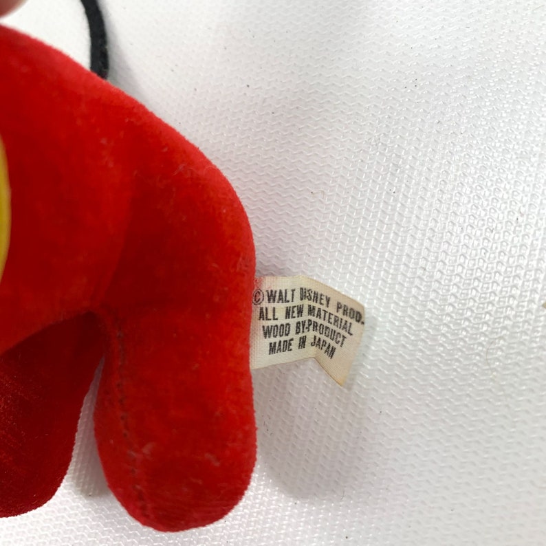 May include: Red plush toy with a yellow face and black eyes. The toy has a tag attached that reads "&copy; WALT DISNEY PROD. ALL NEW MATERIAL WOOD BYPRODUCT MADE IN JAPAN".