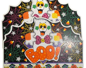 Puede incluir: Un letrero de Halloween plano y troquelado con dos fantasmas blancos de ojos amarillos, parches de lunares naranjas y pajaritas moradas. El fondo muestra telarañas negras, estrellas blancas y caramelos de maíz. La palabra BOO! es naranja, con calabazas debajo.