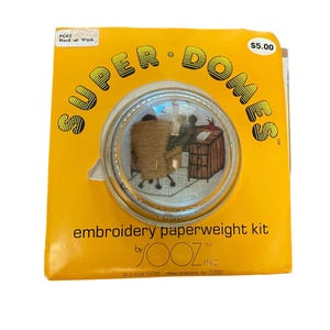 May include: A yellow and white embroidery kit for a paperweight. The kit features a small, clear dome with a stitched image of two people working at a desk. The packaging includes the text "SUPER DOMES" and "Hard at Work".