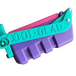 Puede incluir: Una fiambrera de pl&aacute;stico morada y turquesa con las palabras "COOL GEAR" impresas en la parte delantera. La fiambrera tiene una tapa con bisagras y un asa.