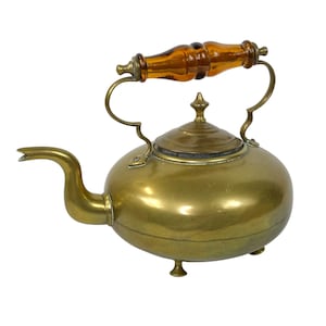 May include: A vintage brass kettle with a rounded body, a curved spout, and a lid with a small finial. The handle is made of brass with a brown, amber-colored glass grip. The kettle has three small feet.