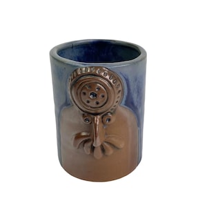 May include: A blue and brown ceramic mug with a face sculpted on the front. The face has a large, round nose and two small eyes. The mug is glazed in a blue and brown color.
