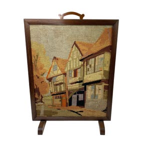 May include: A wooden fire screen with a hand-stitched tapestry depicting a row of colorful houses. The screen has a brown wooden frame and a decorative handle at the top.