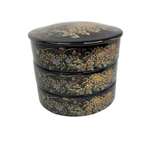 May include: A black and gold three-tiered ceramic serving dish with a floral design. The dish is decorated with chrysanthemums and other flowers.
