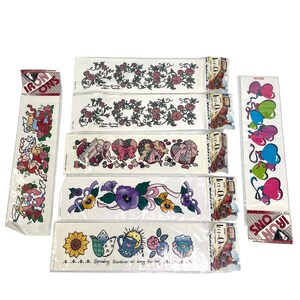 May include: A collection of vintage iron-on transfers featuring floral, heart, and angel designs. The transfers include roses, pansies, sunflowers, and decorative ribbons. The color palette includes pink, purple, blue, and yellow.