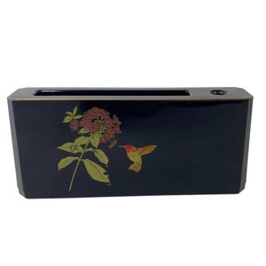 Puede incluir: Una caja negra rectangular con un diseño floral y un colibrí. La caja presenta un fondo oscuro con una ilustración detallada de una flor marrón y un colibrí con acentos rojos y amarillos. La caja tiene una abertura rectangular en la parte superior.