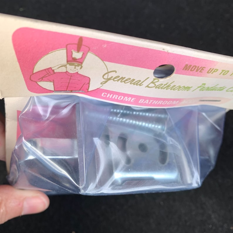 May include: Chrome bathroom hardware set in a clear plastic package. The packaging features a pink and white design with a saluting cartoon figure in a red uniform. The text "General Bathroom Products Co." is visible.