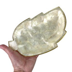 May include: A decorative leaf-shaped dish with a shimmering, pearlescent surface. The dish has a gold-colored metal trim. The leaf-shaped dish is held in a hand against a white background.