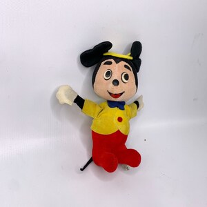 May include: Vintage plush Mickey Mouse toy with black ears, yellow shirt, red pants, and a black tail. The toy has a yellow halo and a blue bow tie.