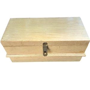 Puede incluir: Una caja rectangular de madera con un acabado en color beige claro y veta natural. La caja cuenta con una tapa con bisagras y un cierre metálico de color latón situado verticalmente en el centro. La base tiene un diseño escalonado.
