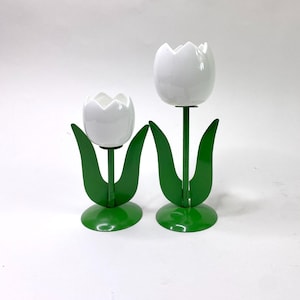 Może przedstawiać: Dwa białe ceramiczne świeczniki w kształcie tulipanów z zielonymi metalowymi łodygami i liśćmi. Świeczniki są różnych rozmiarów.