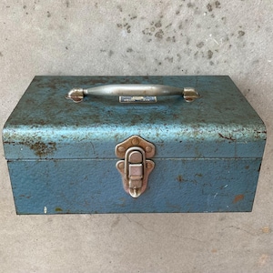 May include: A rectangular, weathered blue metal tool box featuring a silver-toned metal handle on the top and a central metal latch mechanism. The surface shows signs of oxidation and wear, with a small, partially obscured label near the handle.
